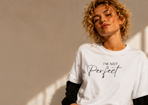 Woman wearing a white sweatshirt with 'I'm not perfect' text, sitting in a sunlit room.